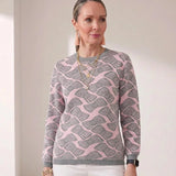 A woman modeling a pink and gray patterned crewneck sweater with white trousers, standing in a brightly lit minimalist room.