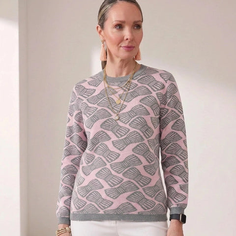 A woman modeling a pink and gray patterned crewneck sweater with white trousers, standing in a brightly lit minimalist room.