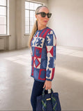 A woman wearing a colorful geometric patterned sweater, dark sunglasses, and dark pants stands in a large, sunlit empty room, holding a navy blue handbag.
